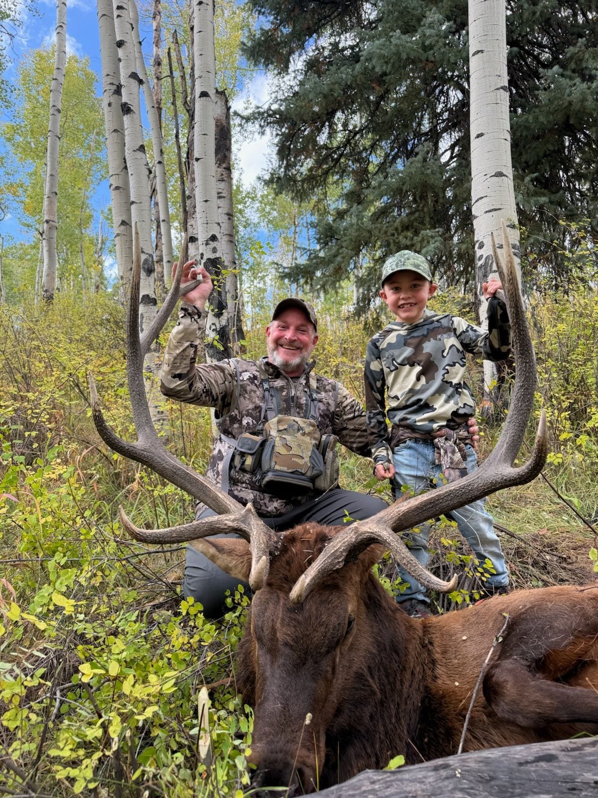 A man and a child pose with a harvested elk in a forest.