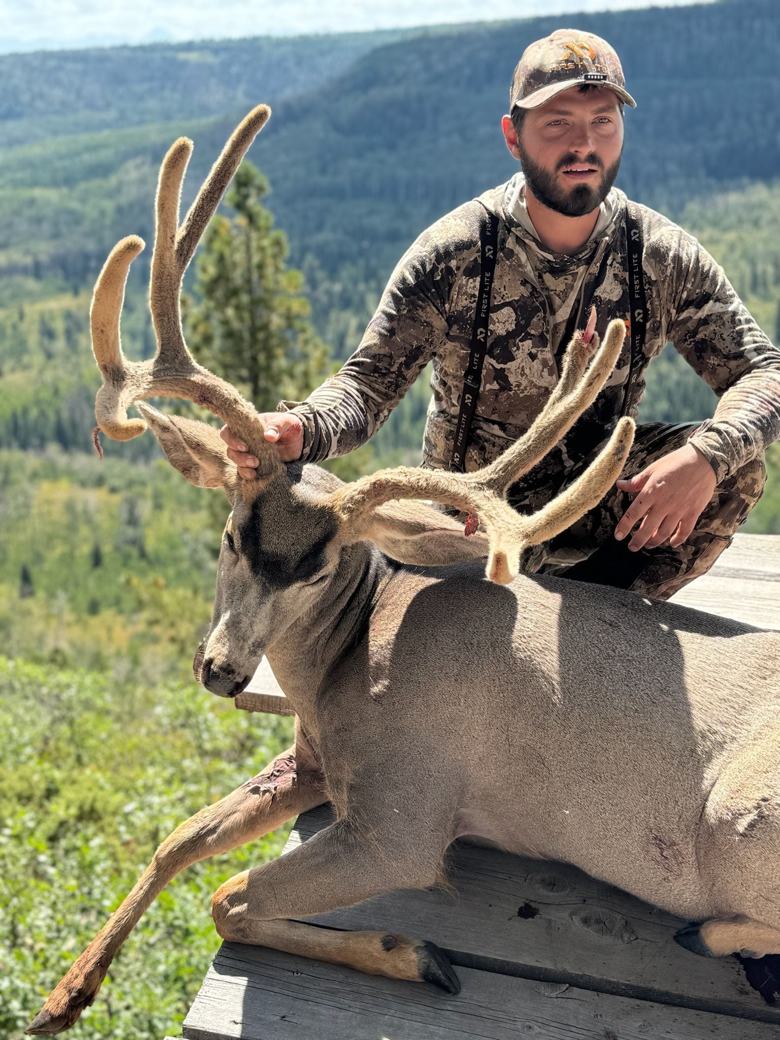 A hunter poses with a harvested mule deer on a wooden platform with a mountainous background.