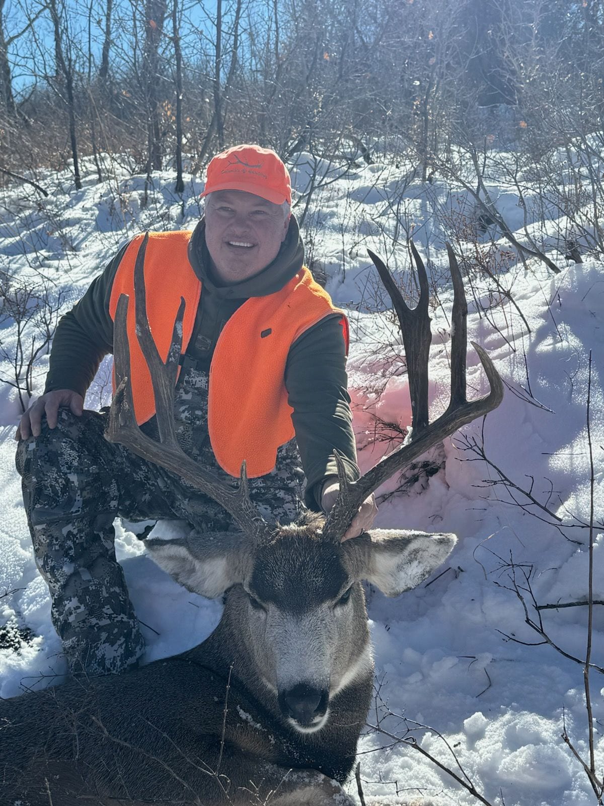 A hunter poses with a harvested mule deer in a snowy, wooded area.