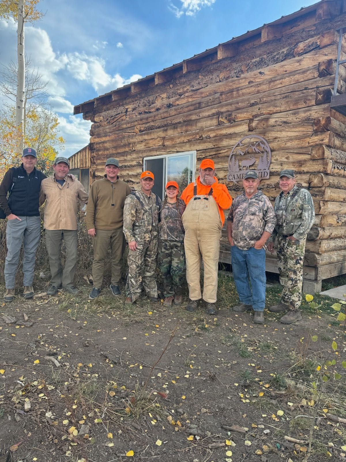A group of people, some in hunting attire, stand in front of a log cabin.