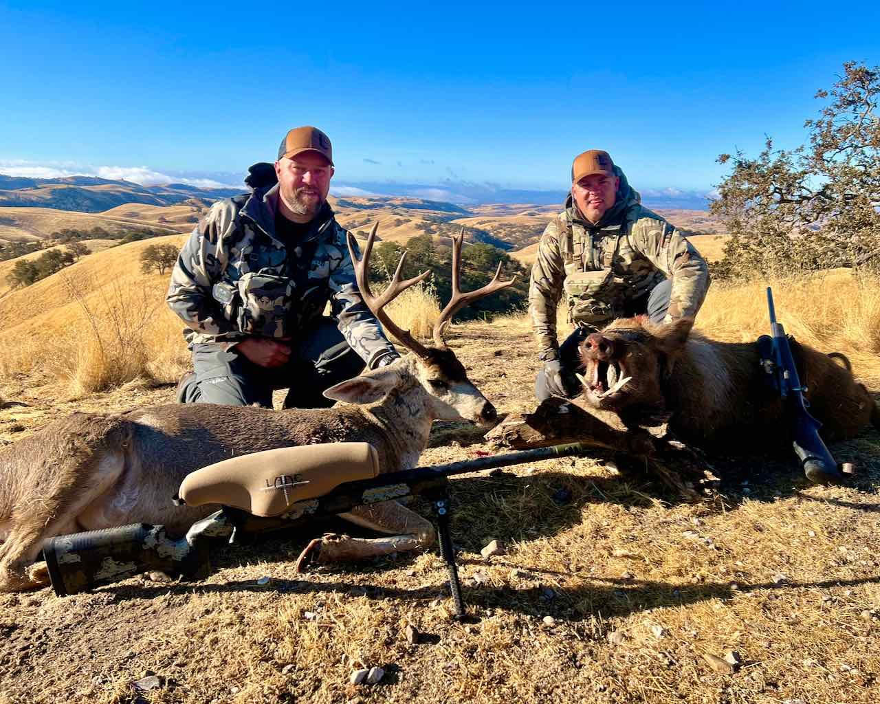 Two hunters pose with a harvested Columbian blacktail deer and a wild boar in a dry, hilly landscape.