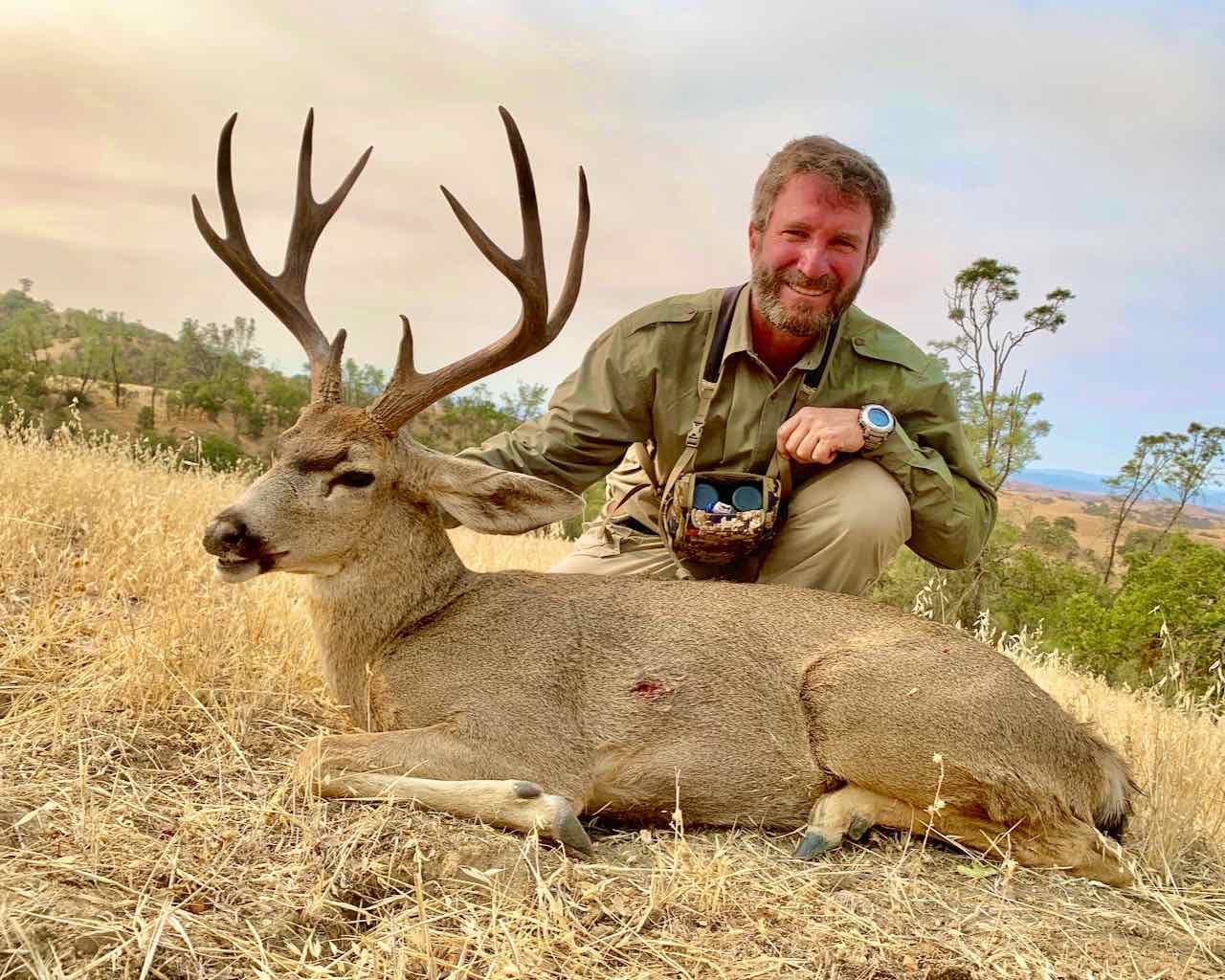 A hunter poses with a harvested Columbian blacktail deer in a field.