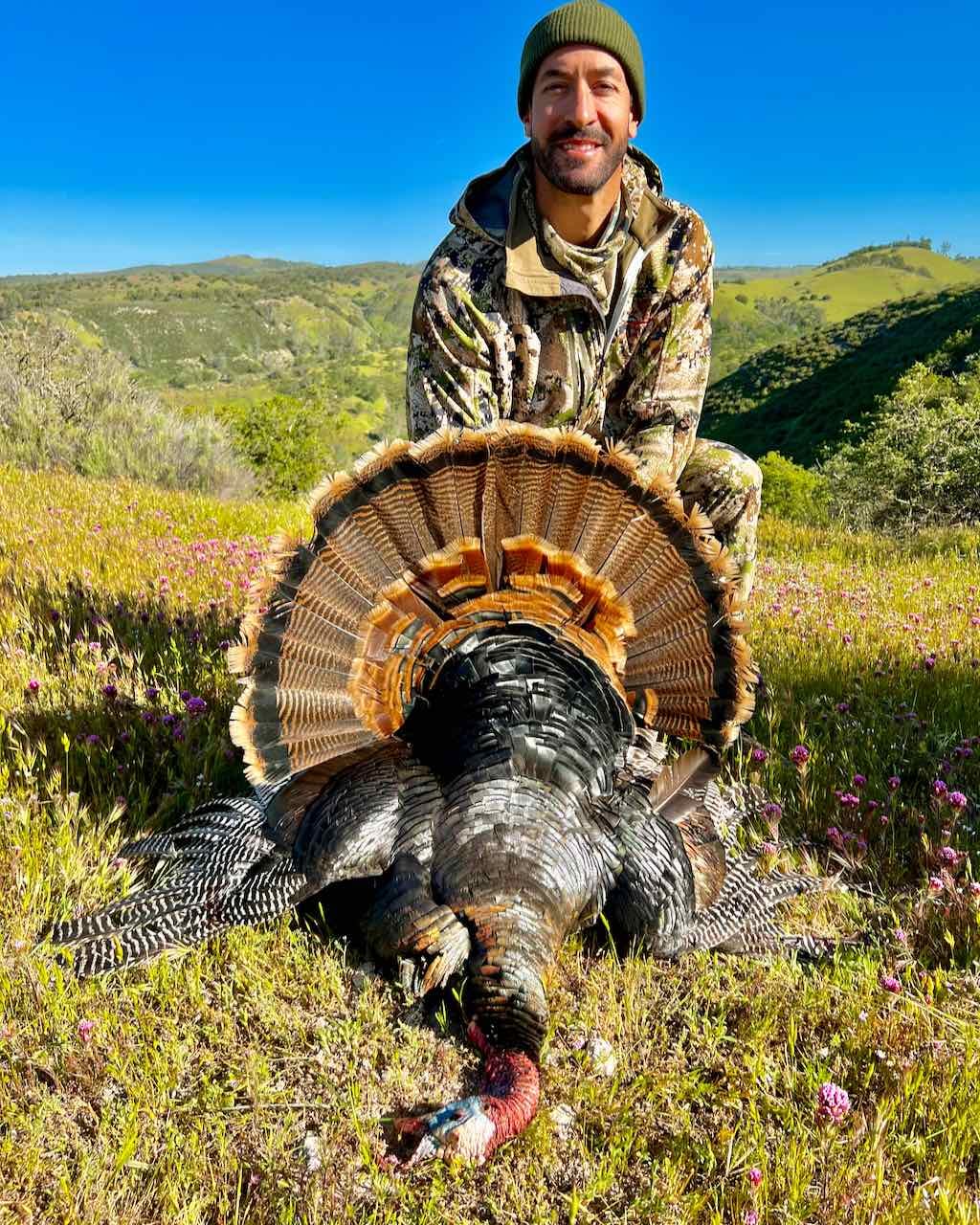 A hunter in camouflage poses with a harvested wild turkey in a grassy, hilly landscape under a clear blue sky.