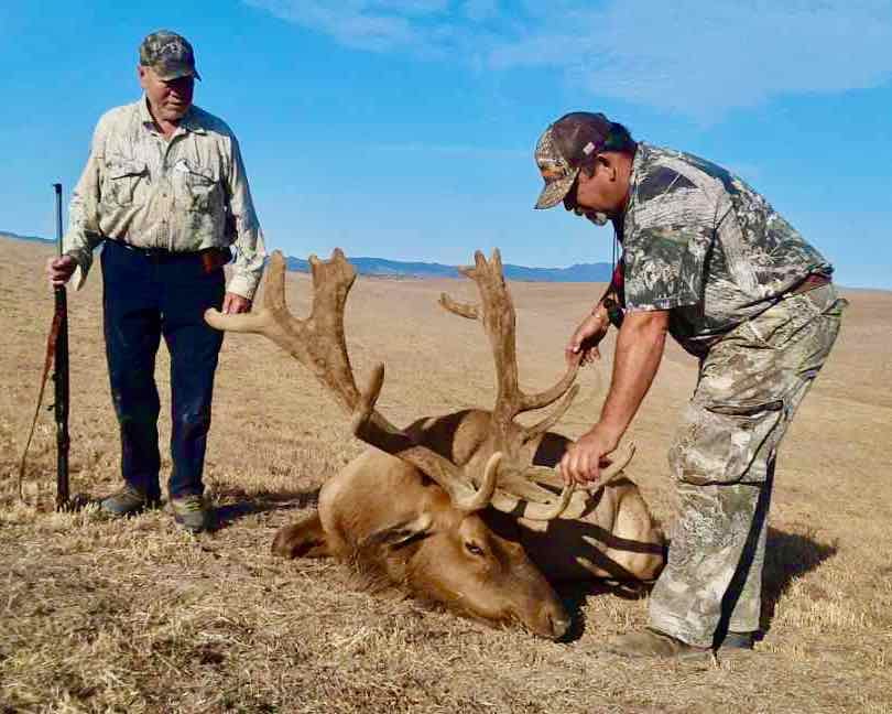 Two hunters pose with a harvested elk in a dry, open field under a clear sky.