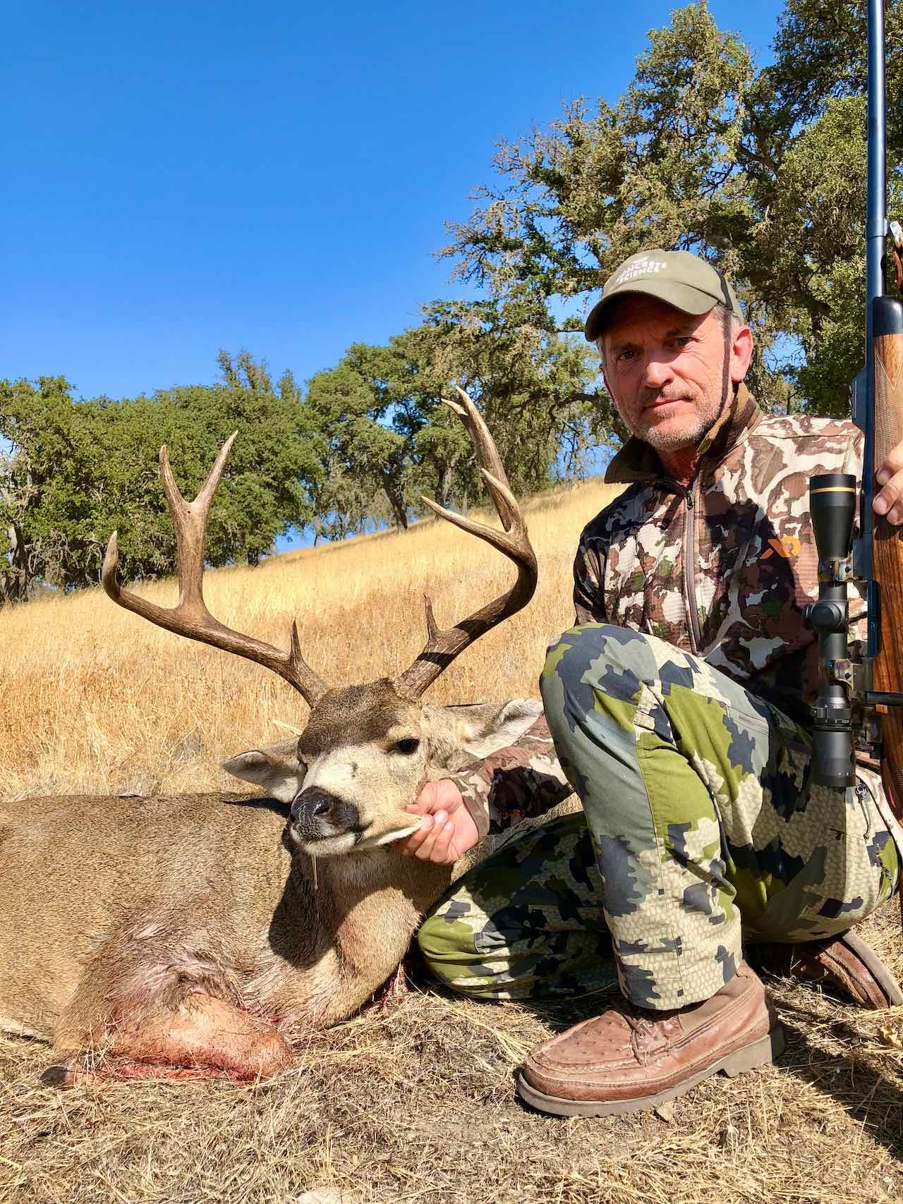A hunter kneels next to a harvested deer with antlers in a grassy field.