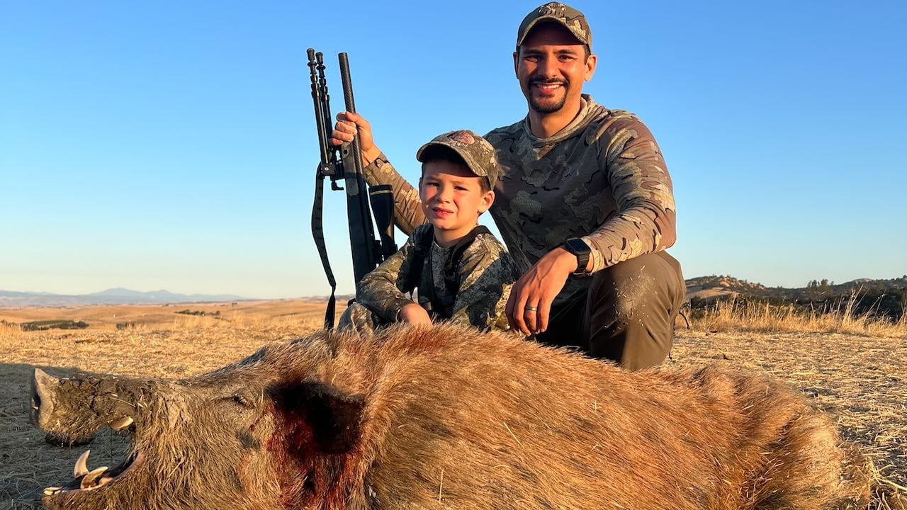 A man and a child pose with a harvested wild boar in a field.