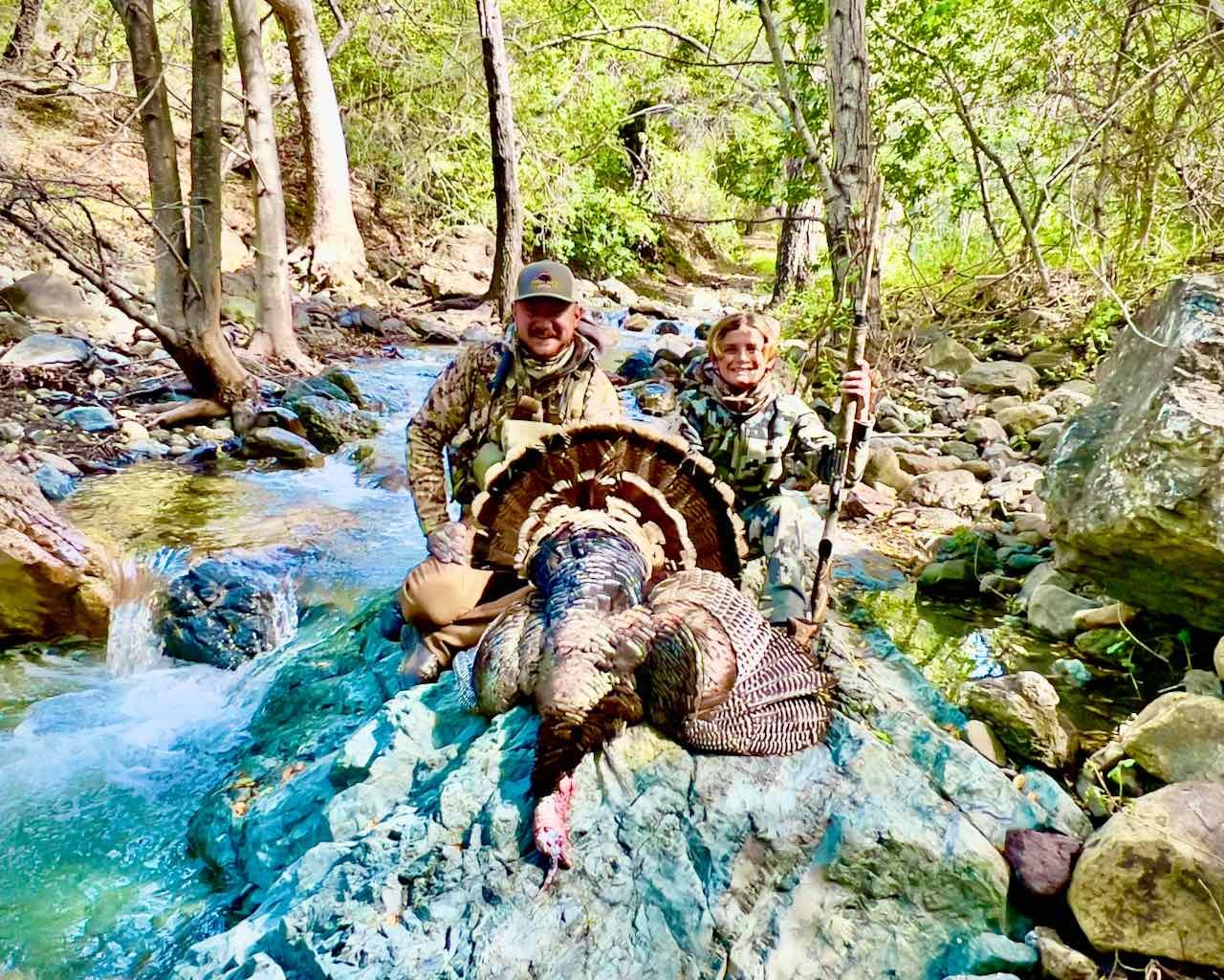 Two hunters, an adult and a child, pose with a harvested wild turkey on rocks next to a stream.
