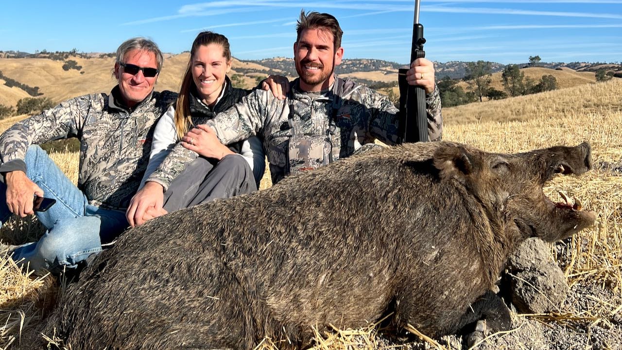 Three hunters pose with a harvested wild boar in a field.