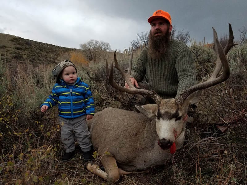 A man and a child pose with a harvested mule deer in a field.