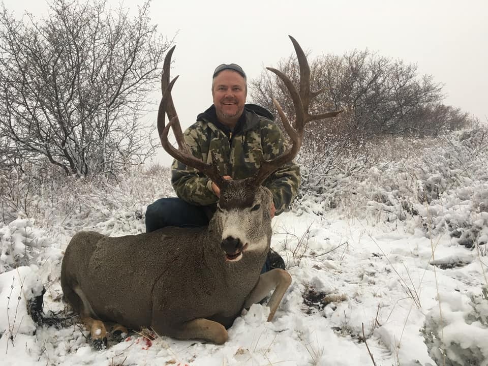 A hunter kneels next to a harvested mule deer in a snowy landscape.