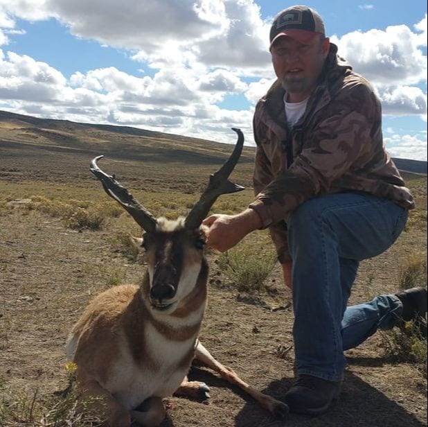 A hunter kneels next to a harvested pronghorn in a field under a cloudy sky.