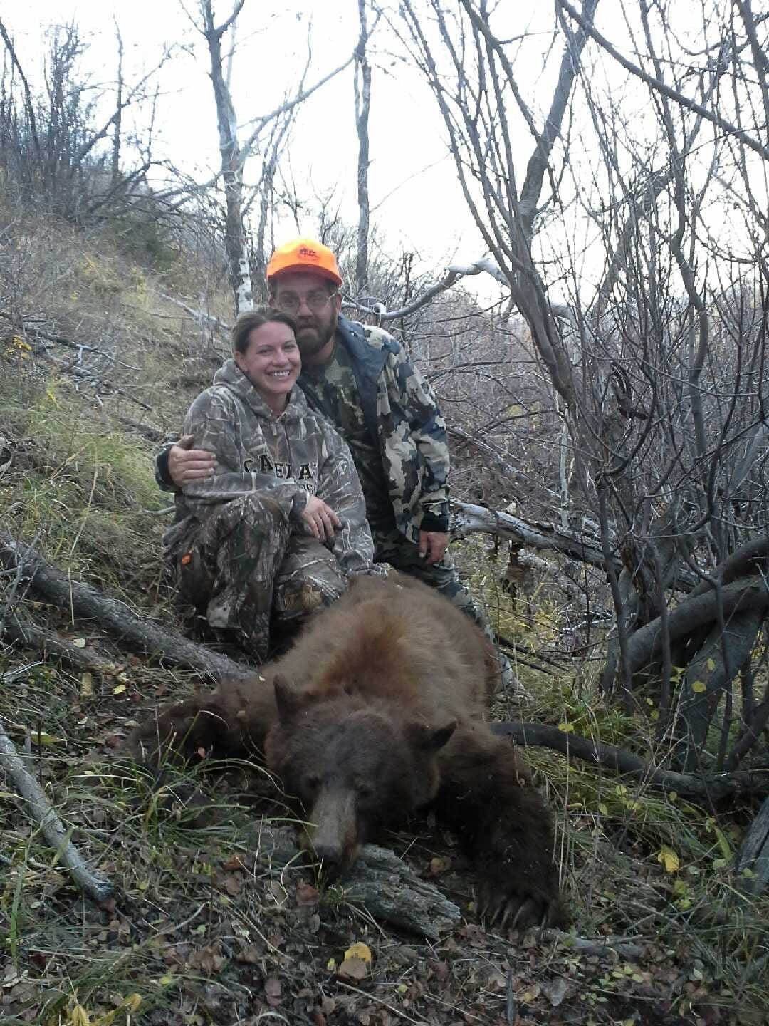 A man and a woman pose with a harvested black bear in a wooded area.
