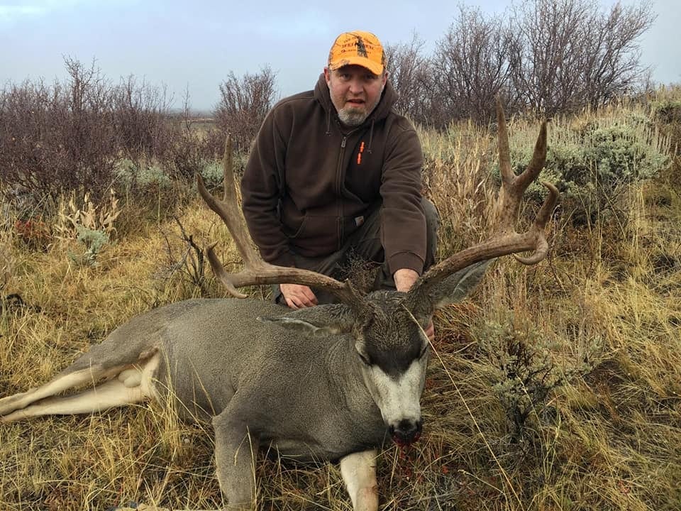 A hunter kneels next to a harvested mule deer in a grassy field.