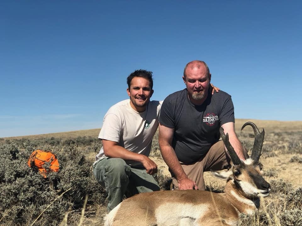 Two men pose with a harvested pronghorn in a field under a clear blue sky.