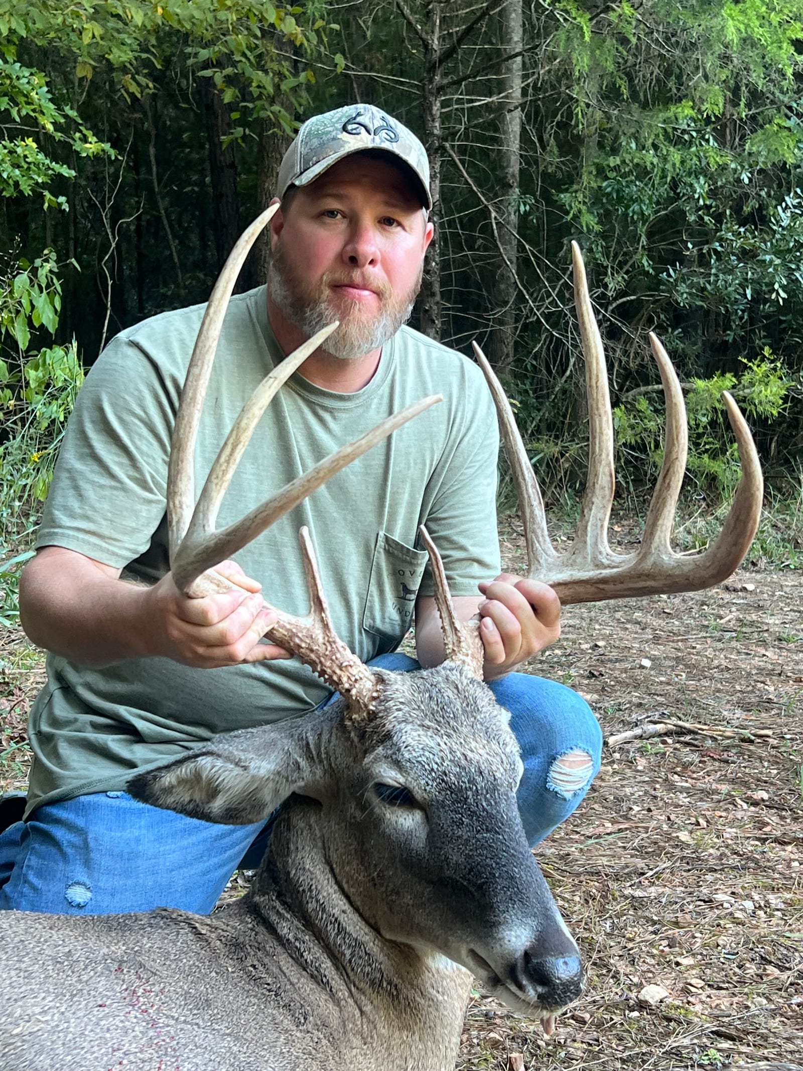 A man in a cap and t-shirt kneels next to a harvested whitetail deer, holding its antlers.