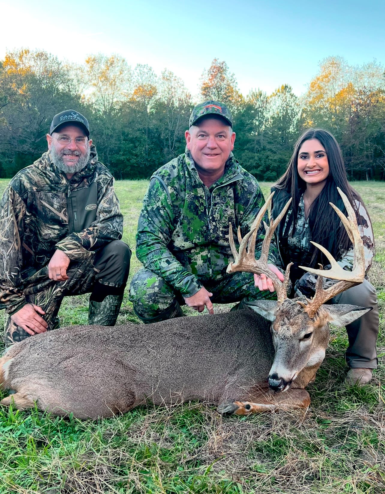 Three people pose with a harvested whitetail deer in a grassy field.