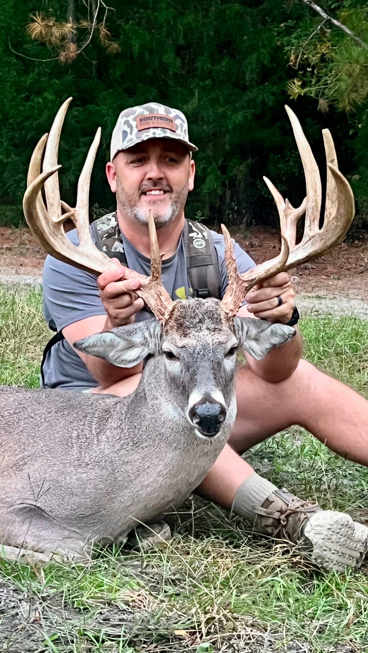 A man in a camouflage hat and vest poses with a harvested whitetail deer.
