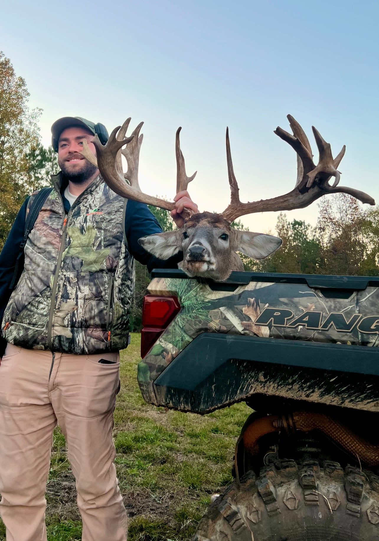A man poses with a harvested whitetail deer in the back of a utility vehicle.