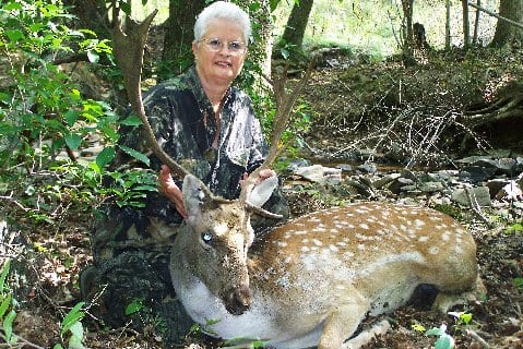 A woman in camouflage kneels next to a harvested fallow deer in a wooded area.
