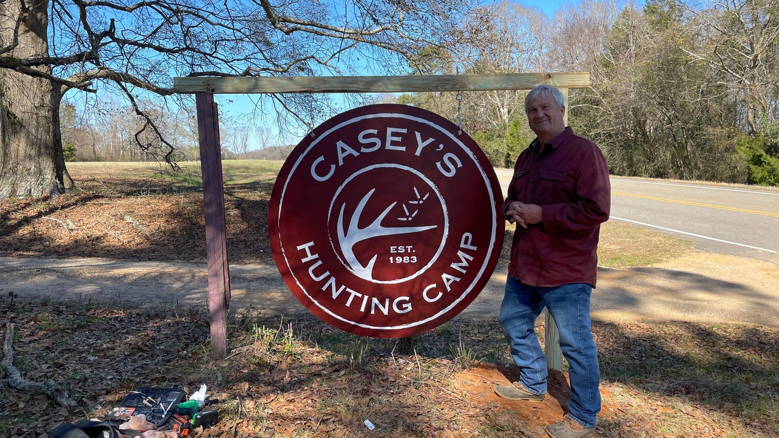 A man stands next to a large circular sign for Casey's Hunting Camp.