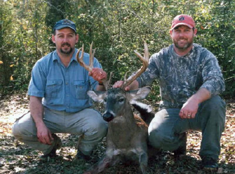 Two hunters pose with a harvested whitetail deer in a wooded area.