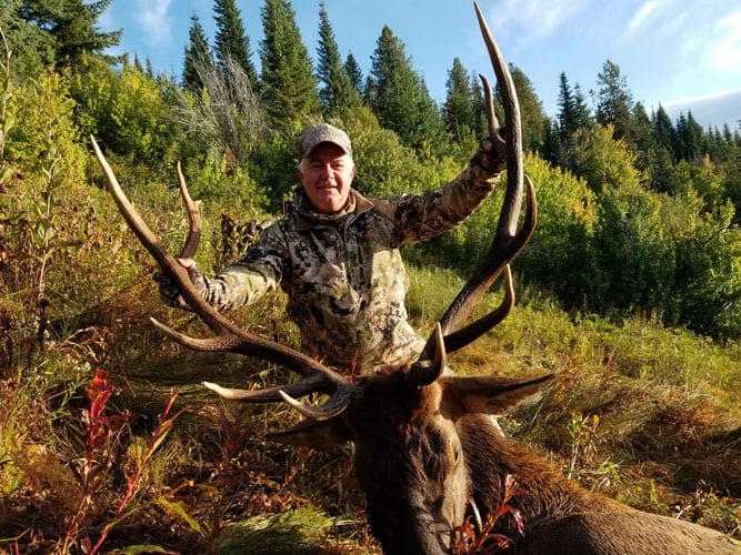A hunter in camouflage poses with a harvested elk in a forest setting.