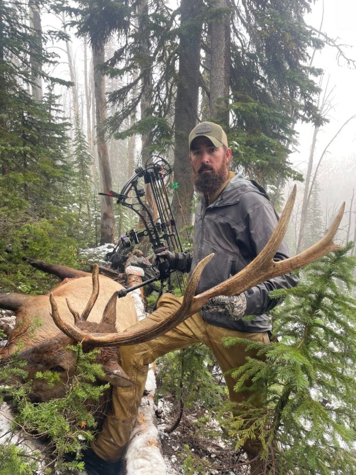 A hunter poses with a harvested elk in a snowy, forested area.