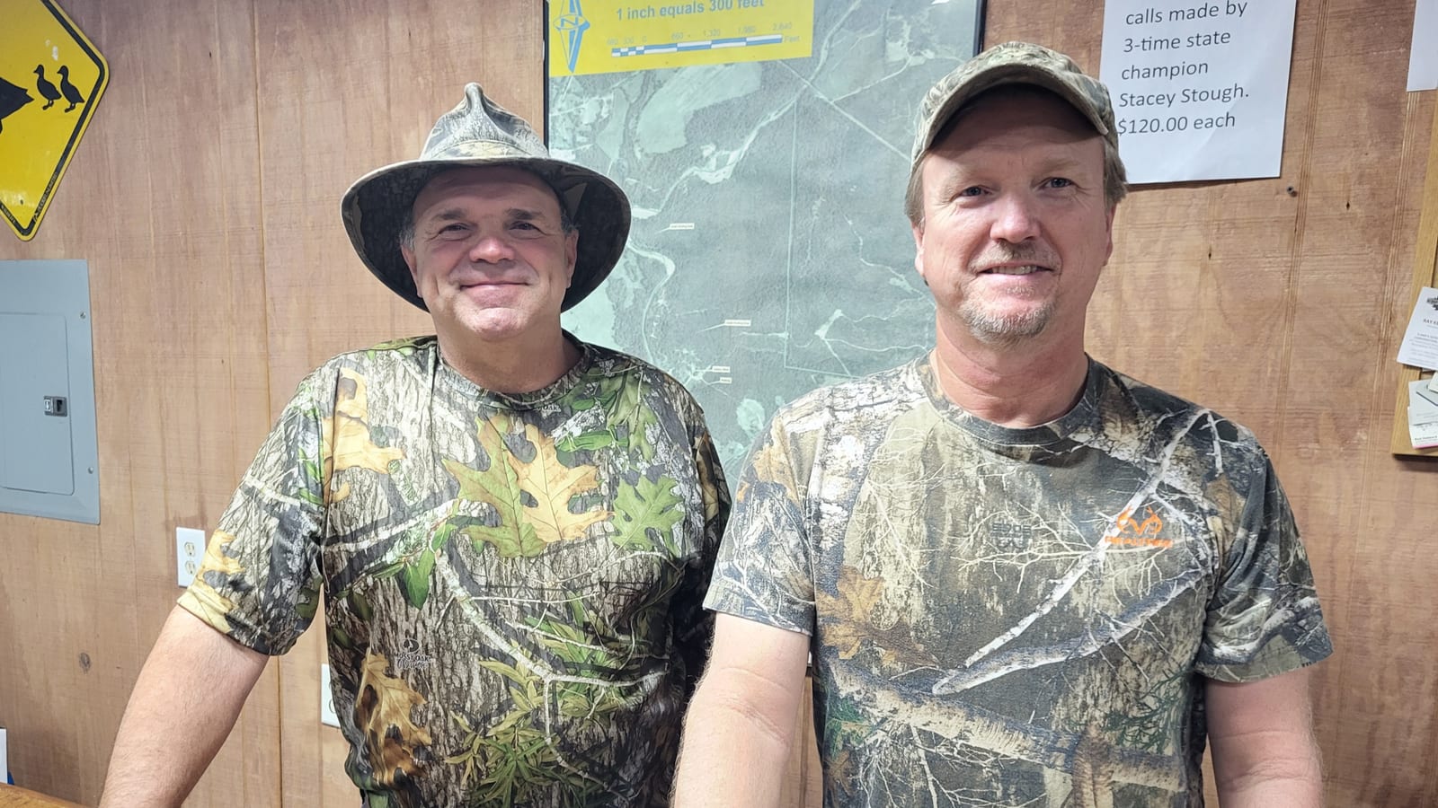 Two men in camouflage shirts stand indoors.
