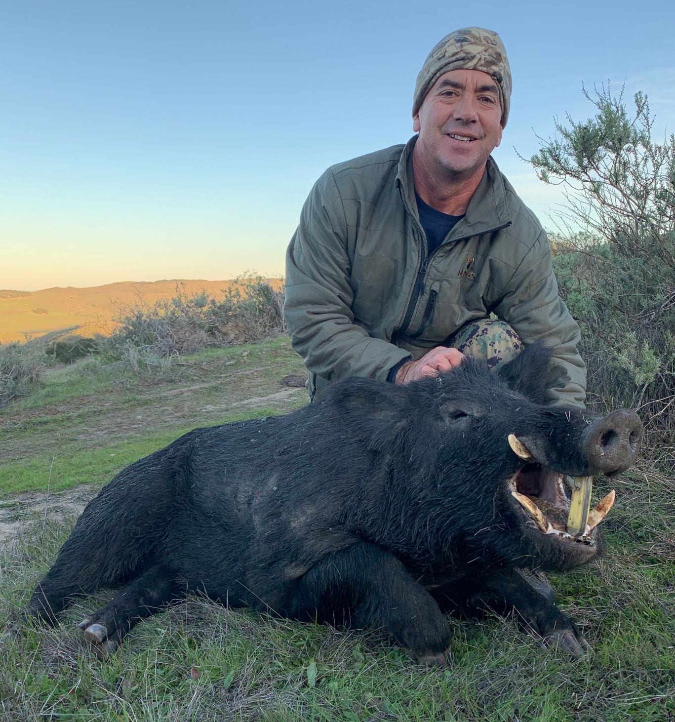 A hunter poses with a harvested wild boar in a grassy field.