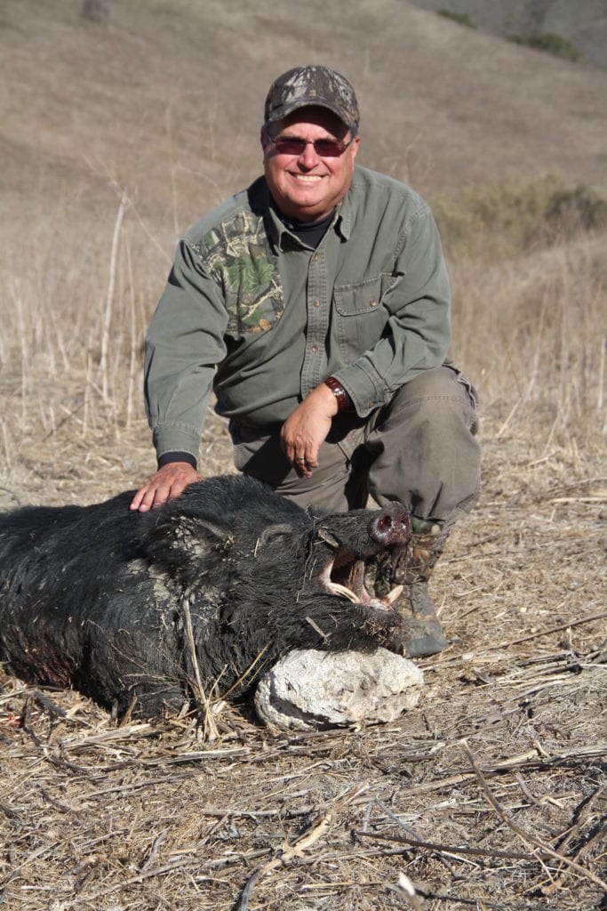 A hunter kneels next to a harvested wild boar in a dry, grassy field.