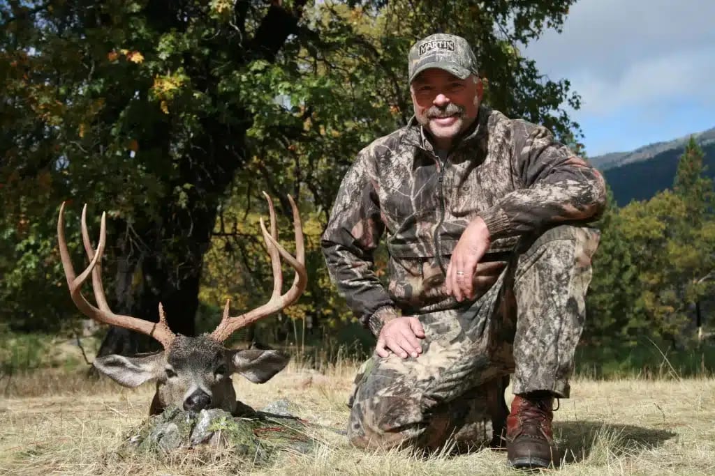 A hunter in camouflage kneels next to a harvested Columbian blacktail deer with large antlers.