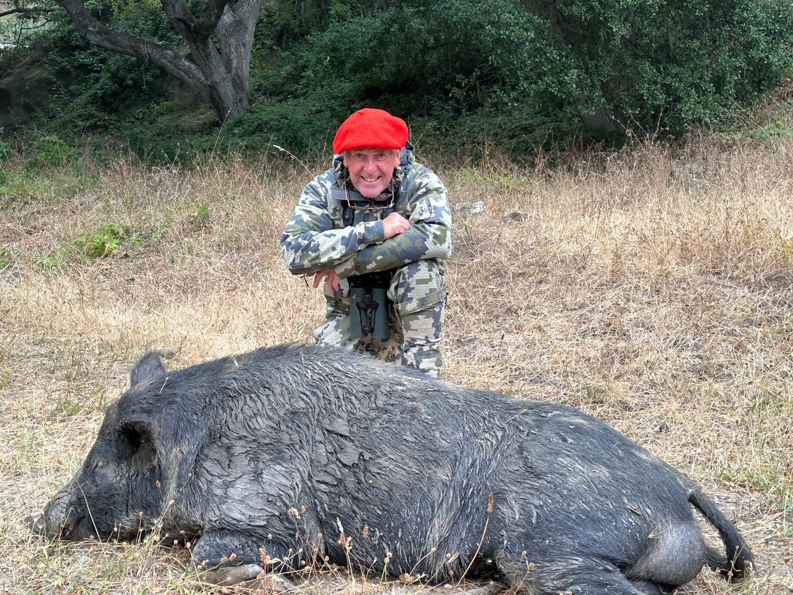 A hunter in camouflage and a red beret kneels behind a harvested wild boar.