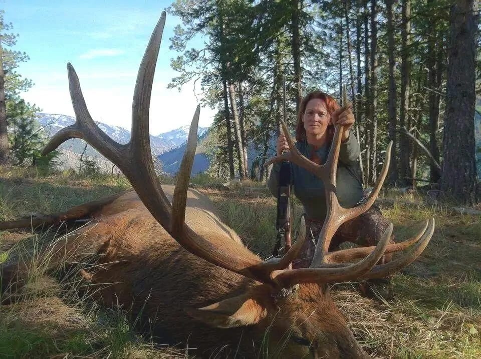 A woman poses with a harvested elk in a forest with mountains in the background.