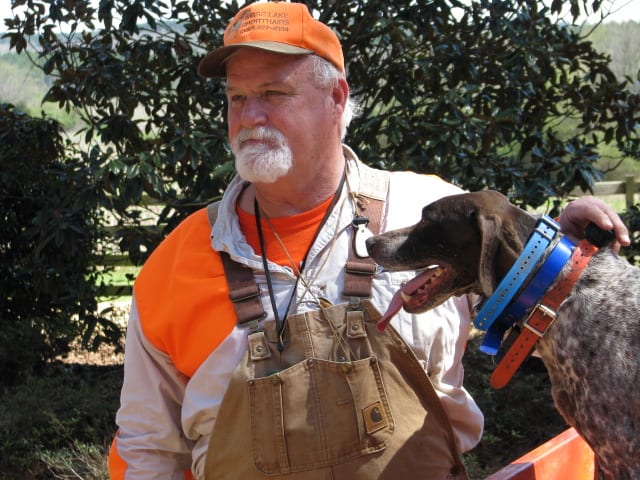 A man in hunting attire stands next to a brown and white speckled hunting dog wearing blue and orange collars.
