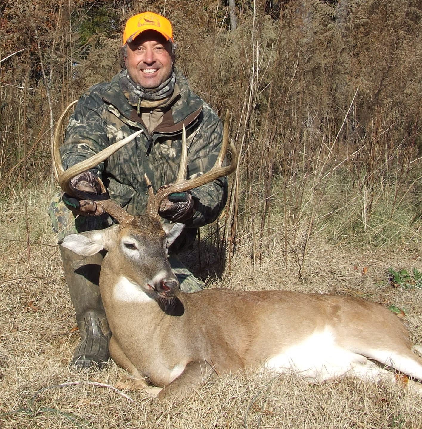 A hunter in camouflage and an orange hat poses with a harvested whitetail deer.
