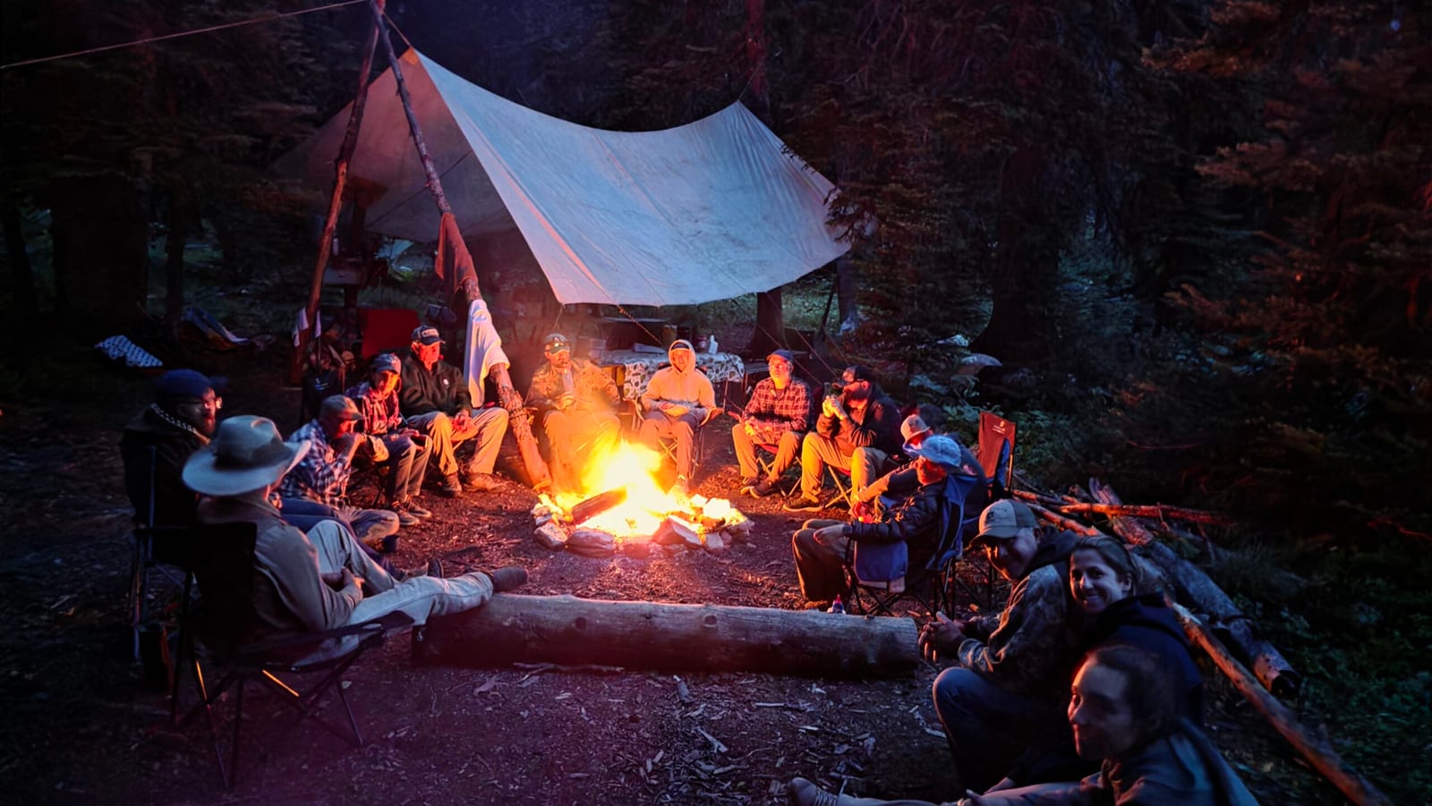 A group of people gathered around a campfire at night under a tarp shelter.