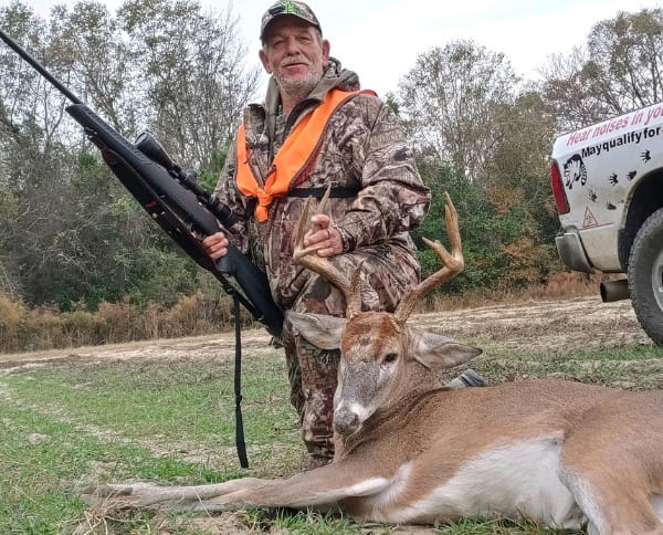 A hunter in camouflage and an orange vest poses with a harvested whitetail deer.