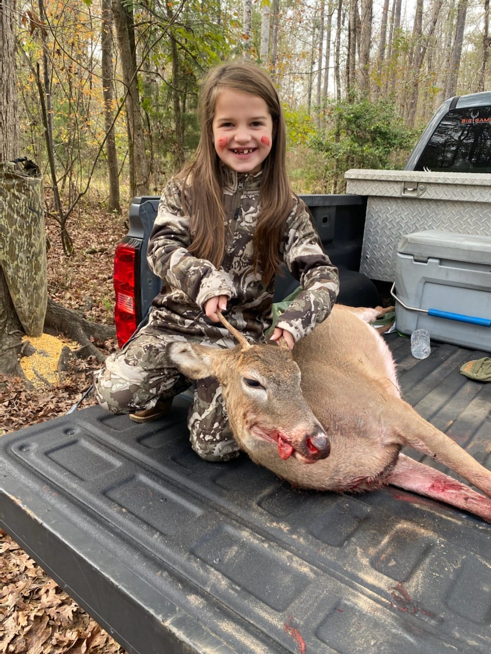 A young girl in camouflage poses with a harvested whitetail deer in the back of a pickup truck.