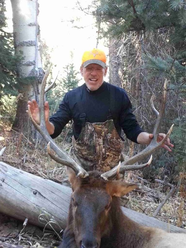 A hunter in a yellow hat and camouflage overalls poses with a harvested elk in a forest.