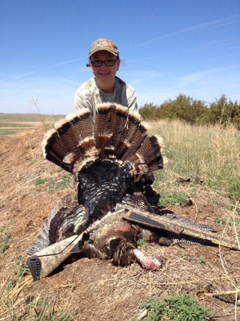 A hunter poses with a harvested wild turkey in a field.