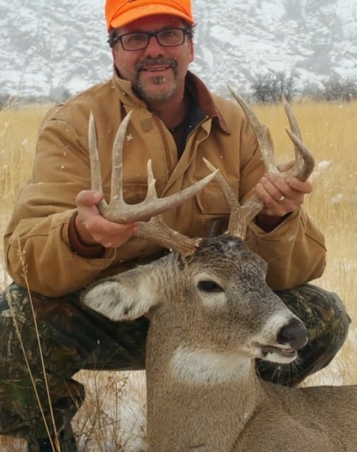 A hunter in an orange hat and brown jacket poses with a harvested whitetail deer in a snowy field.