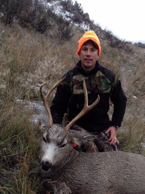 A hunter in an orange hat poses with a harvested mule deer on a snowy hillside.