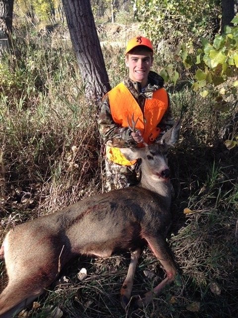 A young hunter in an orange vest and camouflage poses with a harvested mule deer in a wooded area.