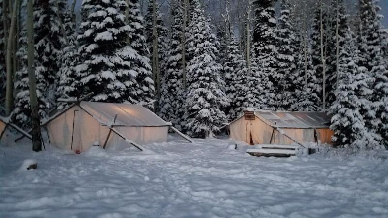Two illuminated canvas tents are set up in a snowy forest.