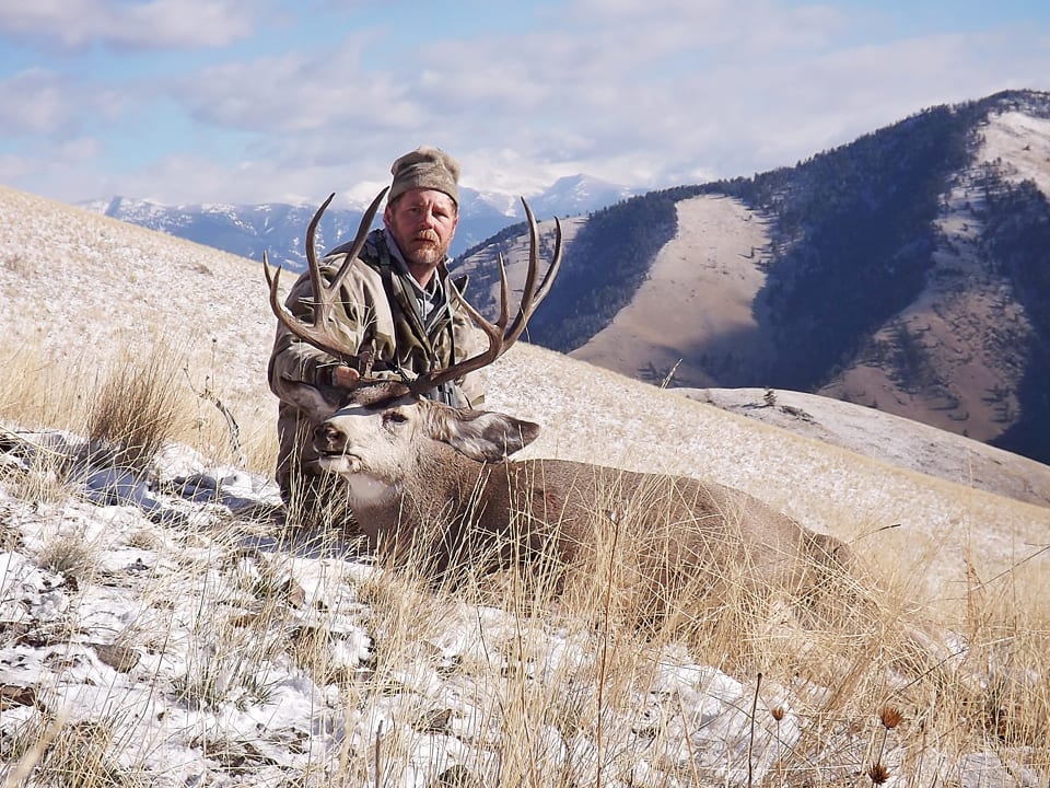 A hunter poses with a harvested mule deer in a snowy, mountainous landscape.