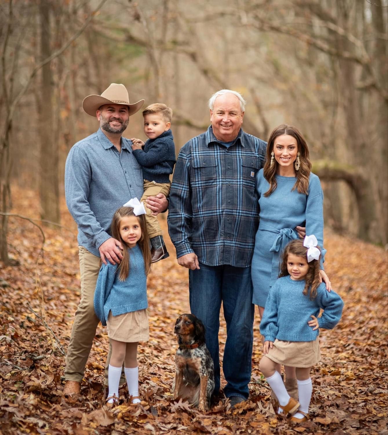 A family with a dog poses for a portrait in a wooded area with fallen leaves.