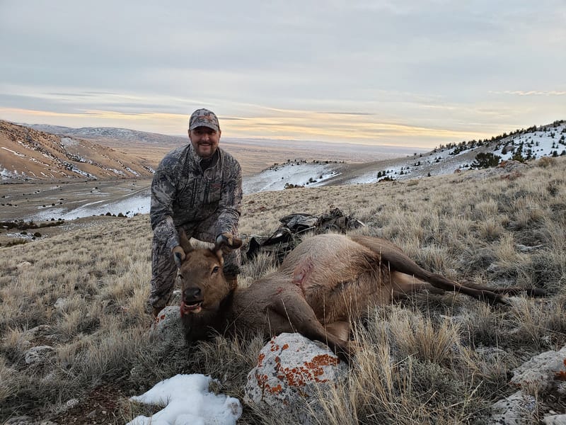 A hunter in camouflage poses with a harvested elk on a snowy, grassy hillside with mountains in the background.