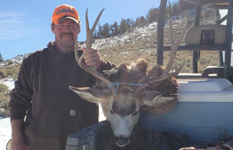A hunter poses with a harvested mule deer in a snowy, mountainous landscape.