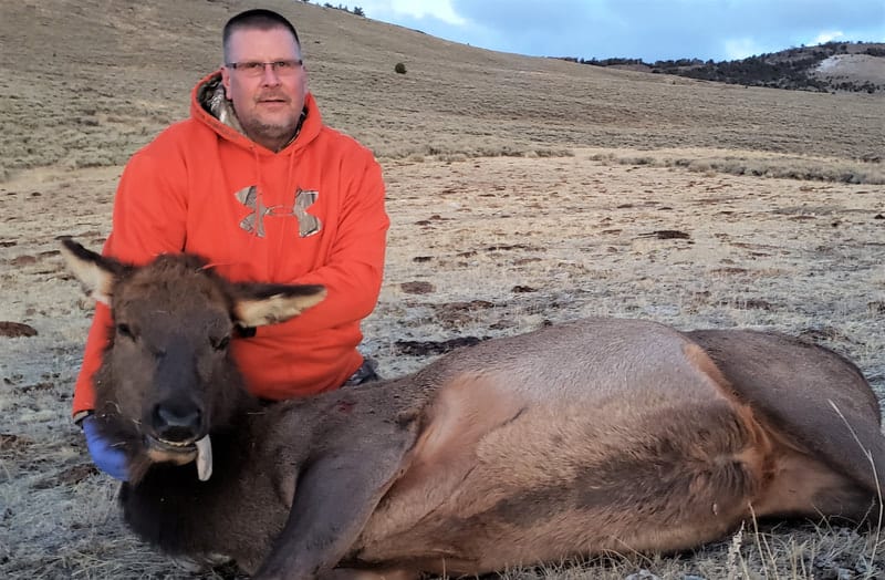 A man in an orange hoodie kneels next to a harvested elk on a dry hillside.