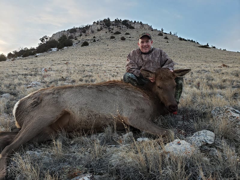 A hunter poses with a harvested elk on a dry, grassy hillside.